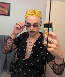 A transfemme goth baddie in fishnets with a button up shirt and thin sunglasses